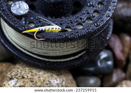 Close up of single trout fly, focus on golden barbed hook with shallow depth of field, on wet fishing reel with blurred out river rocks in background