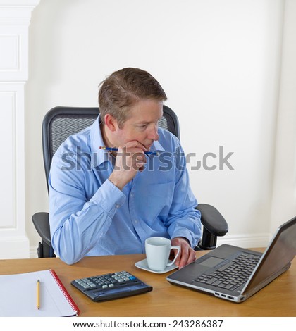 Mature man looking at laptop screen, pencil in hand, while working with white wall in background