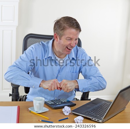 Mature man ripping paper while looking down at desktop with laptop, calculator, broken pencils, wadded paper, coffee cup and notepad on desk. Background is white walls.