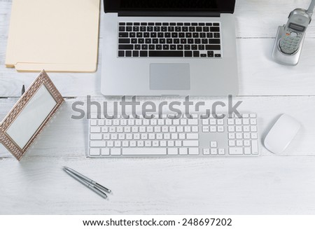 Top view angle of desktop layout for proper use consisting of laptop, keyboard, pens, mouse, picture frame, phone, and work folders on white desk.