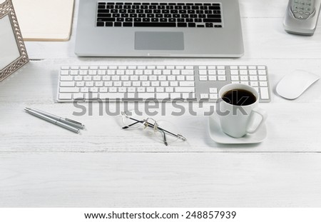 Front view angle of desktop layout for proper use consisting of laptop, keyboard, pens, mouse, picture frame, phone, coffee, reading glasses and work folders on white desk.
