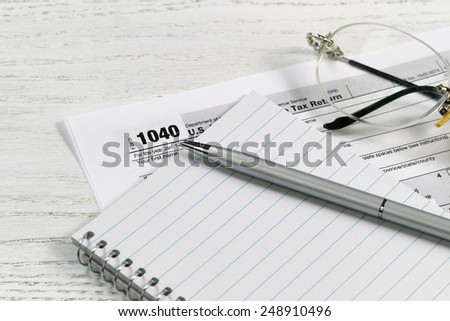 Close up of a silver business pen on top of notepad with tax form and reading glasses in background on white desk. Focus on tip of pen.