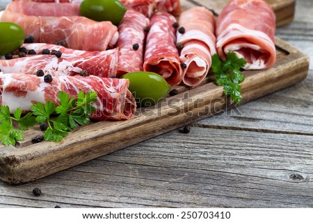 Close up horizontal image of various meats on serving board with ham, pork, beef, parsley, and olives on rustic wood. Focus on side part of serving board and first row of meat.