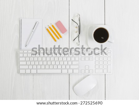 Angled top view image of a business desktop consisting of the following items: keyboard, mouse, pen, pencils, coffee, reading glasses, and note pad on white wood.