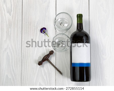 Overhead angled view of a large bottle of red wine, drinking glasses, flower and antique corkscrew on white wooden boards.