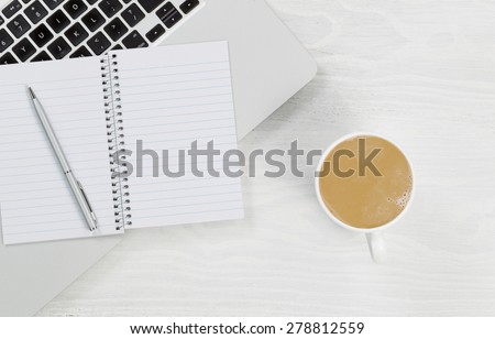 High angle view of laptop, coffee and blank notepad with silver pen on rustic white desktop.