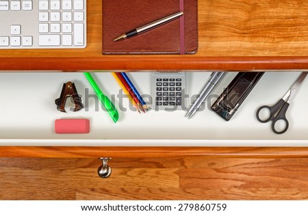 High angle shot of open desk drawer with work items inside. Red oak desktop has computer keyboard, executive notepad and pen. Wooden oak floor underneath desk.
