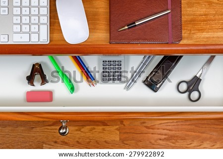 High angle shot of open desk drawer with basic work items inside. Cherry wood desktop has computer keyboard, mouse and executive notepad with pen. Wooden oak floors underneath desk.