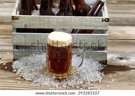 Frosty glass mug of dark beer, focus on front, with vintage crate filled with bottled beer and crushed ice on rustic wooden boards.