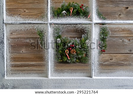Snow covered window with decorative Christmas wreath on rustic wooden boards in background. Focus on window glass and sills.