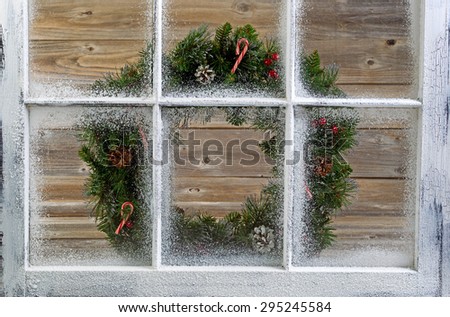 Snow covered window with decorative Christmas wreath on window with rustic wood in background.