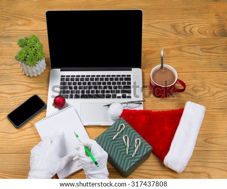 Santa Claus writing his gift list with hot chocolate, computer, present, cap, notepad, pencil, cell phone, reading glasses and plant on desktop. Christmas concept of Santa Claus office.