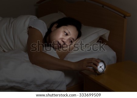 Woman with insomnia touching alarm clock while eyes open. Select light and focus on woman and clock with darker background for night time concept.