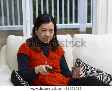 Asian woman sitting on white leather couch with coffee mug and tv remote in hands