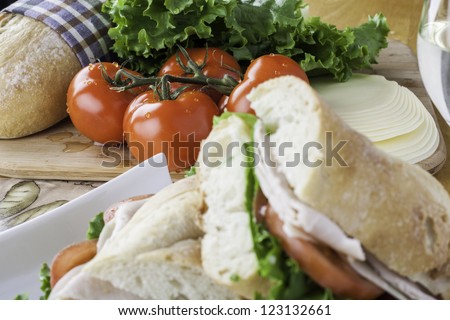 Turkey sub on rustic bread with tomato, bread, lettuce and cheese in the background with the sandwich in the foreground out of focus