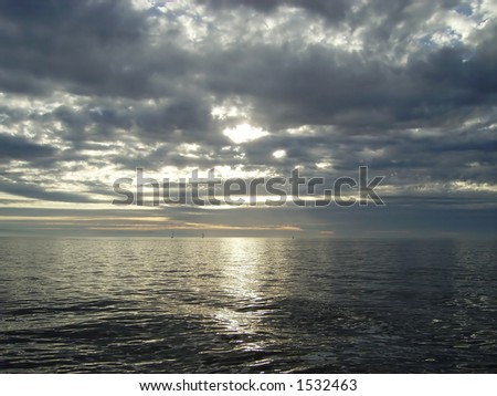 Clouds pass by overhead and cover the sun, which is piercing through and lighting the ocean, especially at the horizon.
