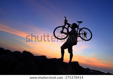 Silhouette of mighty man in action lifting bike on his shoulder on rock mountain with sunrise twilight background. Symbol of relax, success and touring.