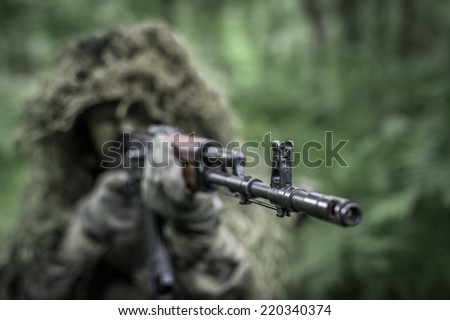 Special forces soldier in heavy camouflage hidden in forest, aiming with assault rifle - focus on barrel