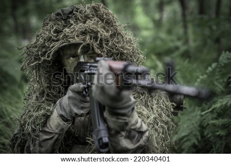 Special forces soldier in heavy camouflage hidden in forest, aiming with assault rifle - focus on eyes