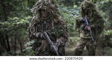 Two special forces soldiers on patrol in deep forest. Operators dressed in ghillie suits, holding assault rifles.