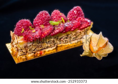 Slice of a tart with fresh raspberries, chopped almond and pistachios on top, golden berry on the foreground.