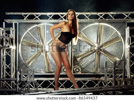 very sexy and beautiful top model on a stage posing with a huge turbo ventilator behind her were the smoke is coming and lights are coming trough