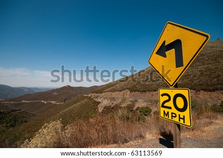Long winding tarmac road in California heading towards Napa Valley, clear blue sky and a dominant bright yellow road sign showing a 20 MPH speed limit approaching a left handed corner