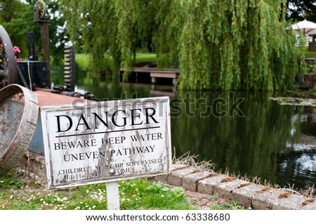 A weather sign beside a river that reads - danger beware deep water uneven pathway, children must be supervised at all times. There is machinery in the foreground and a Willow tree and jetty behind