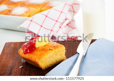 A piece of butter cake with butter and strawberry jam on a wooden plate. Cake in the pan and bottle of milk in the background