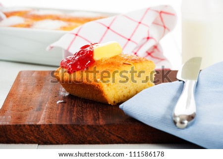 A piece of butter cake with butter and strawberry jam on a wooden plate. Cake in the pan and bottle of milk in the background