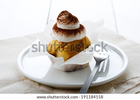 vanilla cupcakes with whipped cream and cocoa powder. White plate, cream color napkin and white wood table