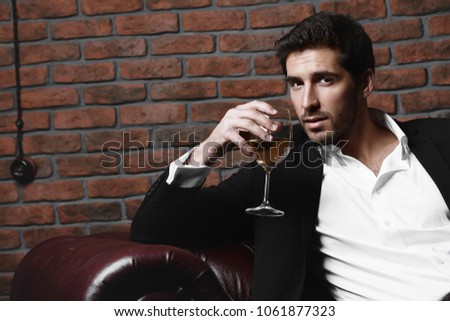 Portrait of a sexy handsome man drinking wine on a leather sofa. Luxurious lifestyle. Fashion shot. Men's clothing and accessories.