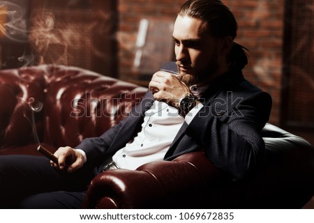 Imposing young man sitting on a leather sofa, drinks whiskey and smokes a cigar in a modern interior. Men\'s beauty, fashion.