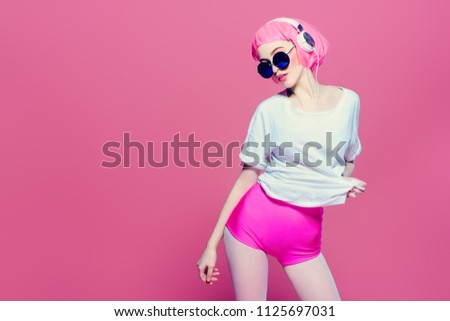 Trendy girl with pink hair wearing sunglasses enjoys the music on headphones. Pink background. Youth style, leisure.