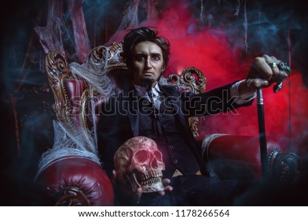 Halloween. Frightening gloomy man in a black tailcoat sits in an armchair with a skull in an old abandoned castle. The Dark Lord. Vampire man.