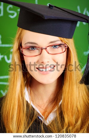 Portrait of a graduating student girl in an academic gown standing at the classroom.