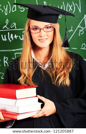 Graduating student girl in an academic gown standing with books at the classroom.