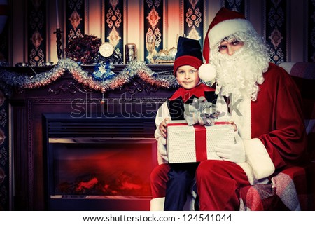 Santa Claus sitting with a little cute boy elf near the fireplace.