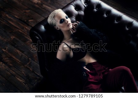 Seductive girl in the black bra, red pants and  fur jacket lying on a leather sofa. Luxurious lifestyle. Fashion, beauty. Studio shot.