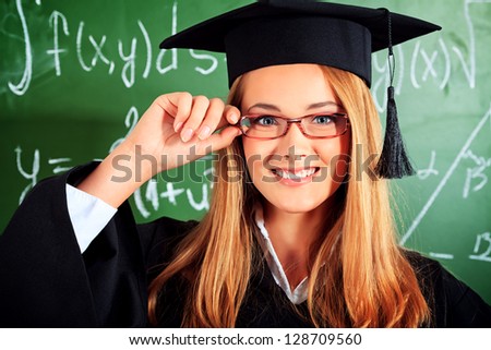 Portrait of a graduating student girl in an academic gown standing at the classroom.