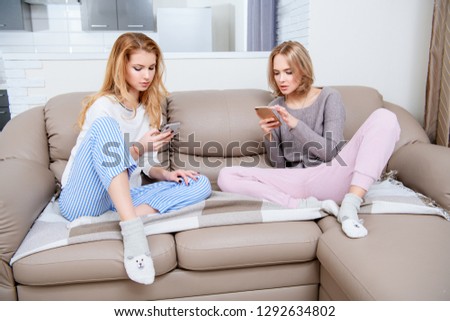 Two girlfriends are using smartphones in home clothes on the couch in the interior. Gadgets, home, casual style.