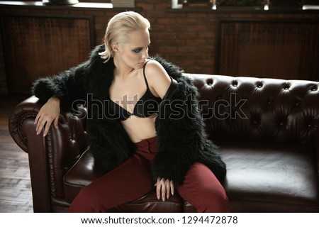 Seductive girl in the black bra, red pants and  fur jacket sitting on a leather sofa. Luxurious lifestyle. Fashion, beauty. Studio shot.