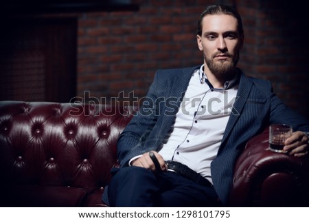Imposing young man sitting on a leather sofa, drinks whiskey and smokes a cigar in a modern interior. Men\'s beauty, fashion.