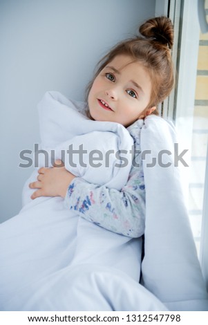 A little girl sittiing on a windowsill on a pillow covered with a blanket. Childhood. Kid\'s fashion.