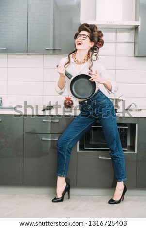 An attractive girl in everyday clothes and curlers stands in the kitchen set with a frying pan. Fashion home shot. Full length portrait.