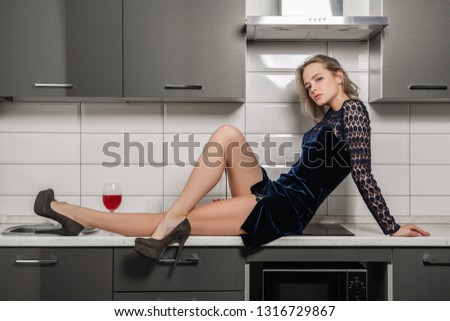 Fashion shot. Beautiful female model with short hair is posing in cocktail dress sitting on the countertop of the kitchen set. Beauty, fashion.