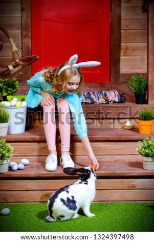 Happy little girl with bunny sitting on the porch near the wooden house. Easter holiday. Rural style, easter decoration.