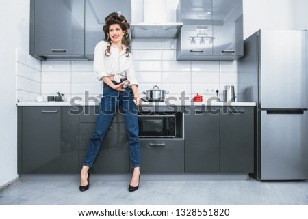 Beautiful girl is posing in everyday clothes with curlers in her hair in the kitchen. Fashion home shot. Pin-up style.