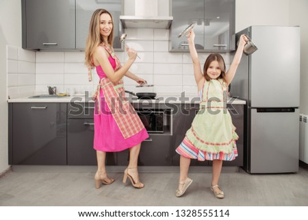 A full length portrait of mother and daughter in aprons with pots in the kitchen.