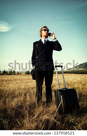 Handsome business man standing in a field with his suitcase and cell phone.